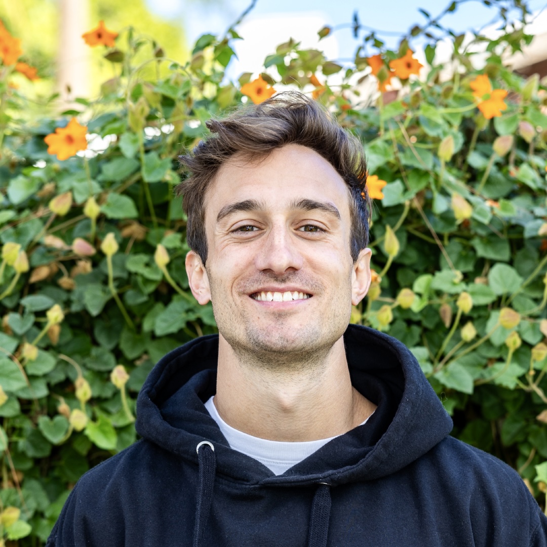 Young man smiling with sweater on and surrounded by flowers and leaves